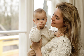 Mother and her little baby son wearing warm sweaters standing by the window