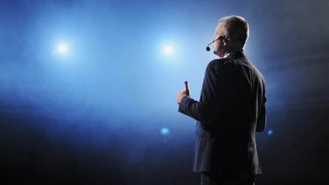 Showman. Middle Age Male entertainer, presenter or actor on stage. Arms to sides, smoke on background of spotlight. Rear view portrait of a male public speaker speaking at the microphone, pointing