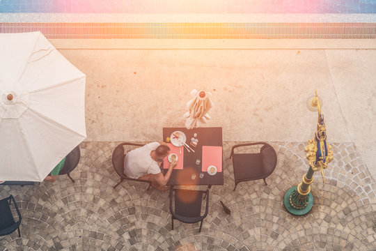 A man sits at a table near the pool, drinking coffee