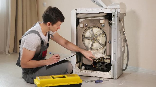 young serviceman checks broken washing machine loader with open back side panel on floor in room close view