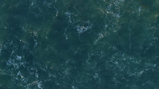 Turquoise ocean waters waving with white foam lit by golden light of setting sun. Aerial top down shot, 4K