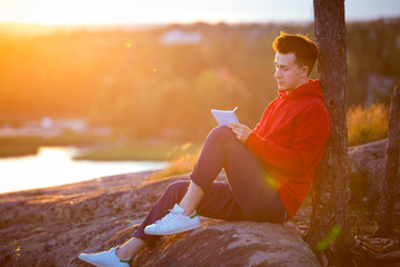  Young man in hoodie sitting on top of the rock, writing in the notebook. Beautiful sunset...