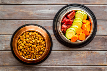 Healthy feed for cats and dogs. Bowls full of dry feed, meat and vegetables on dark wooden background top-down copy space