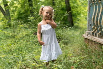  beautiful little girl in a white dress stands near the arbor among green grass. summer vacation in the village.