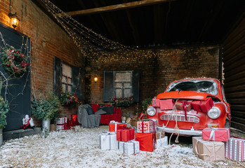 New Year and magic Christmas decorated exterior with retro red car with many gifts and festive lights and garlands outside of house with sofa
