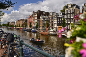 Fototapeta premium Amsterdam,Holland,August 2019. Enchanting view of a canals of the historic center. The typical houses overlook the water, boats moored along the canal. Planters on railings and bikes. Sunny day.