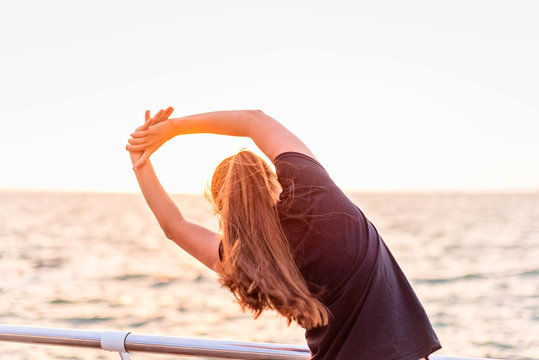 Back young sports girl with long brunett hair in sportswear doing stretching exercises sundial salutation on promenade on the background of the sea at sunrise