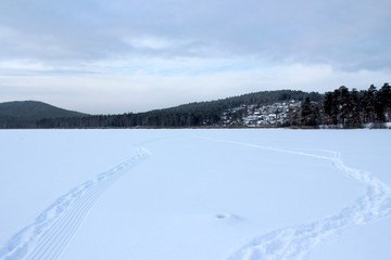 Fototapeta na wymiar Winter landscape on the lake, forest and country houses on the hills in the background. Snow trail and trails extending into the distance to small houses. White silence, snow and sky