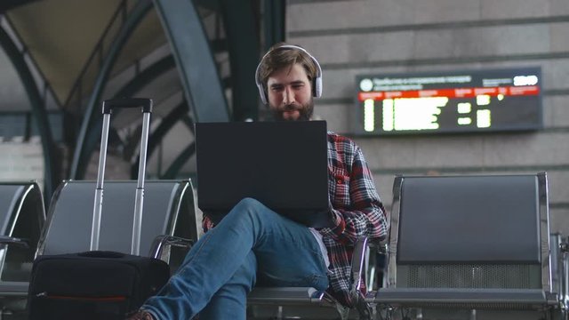 Hipster tourist with headphones and laptop witing fr train at railway station