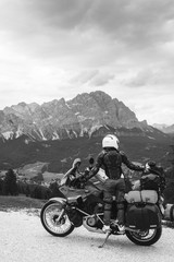 Woman biker with adventure touring motorcycle in full equipment on dirt road, "Monte Cristallo" mountains on background, tourism travel concept, vertical photo Black and white. Cortina Ampezzo, Italy