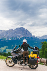 Woman biker with adventure touring motorcycle in full equipment on dirt road, "Monte Cristallo" mountains on background, tourism travel concept, vertical photo, sunny day. Cortina Ampezzo, Italy