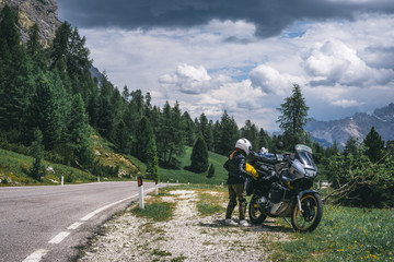 Biker lady with adventure touring motorcycle in full equipment on side of the road, Mountains and forest with rainy clouds on background, tourism concept, copy space summer day, Italy