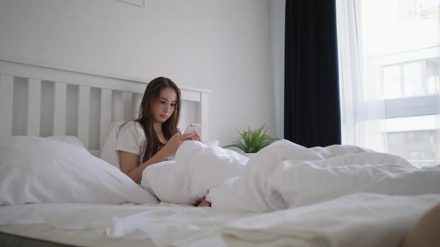 female is lying in bed in morning and looking at screen of her cell phone