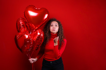 Fototapeta na wymiar Studio portrait of young woman with dark skin and long curly hair wearing knitted turtle neck sweater over the festive red wall with heart shaped balloon. Close up, isolated background, copy space.