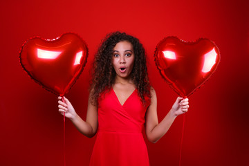 Fototapeta na wymiar Studio portrait of young woman with dark skin and long curly hair wearing sexy dress over the festive red wall with heart shaped balloon. Close up, isolated background, copy space.