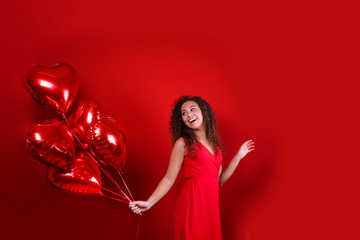 Studio portrait of young woman with dark skin and long curly hair wearing sexy dress over the festive red wall with heart shaped balloon. Close up, isolated background, copy space.