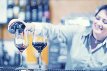 Bartender woman pours red wine into two wineglass to a client in the hotel bar. Shelves with bottles of alcohol in the background. The concept of service. 