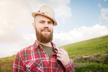 Portrait of a bearded happy smiling traveler hipster with a backpack in a plaid shirt and a hat next to an unknown car stands on the road at sunset in the mountains. Happy and confident travel concept