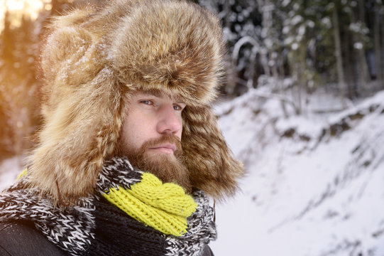 Portrait Of An Attractive Manly Man With A Mustache And Beard In A Winter Forest. Stylish Tourist In A Fur Hat And A Bright Scarf