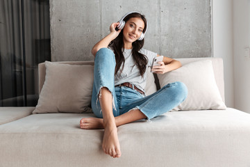 Image of woman in headphones holding smartphone while sitting on sofa