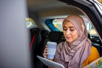 Busy young arabian woman with hijab on head sitting in taxi cab, typing on laptop keypad and talking on smartphone. Portrait of a young, attractive Muslim woman wearing a hijab commuting in a car.