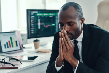 Tired young African man in formalwear keeping hands clasped while working in the office