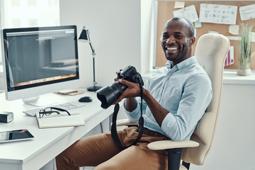 Handsome young African man holding digital camera and smiling while working in the modern office