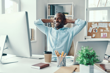 Handsome young African man in shirt keeping hands behind head and smiling while working in the office