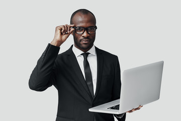 Serious young African man in formalwear working using computer while standing against grey background