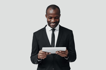 Charming young African man in formalwear working using digital tablet while standing against grey background