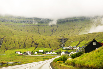 Faroese village with traditional wooden houses under green mountain with a small waterfall and great clouds.