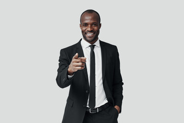 Handsome young African man in formalwear pointing you and smiling while standing against grey background