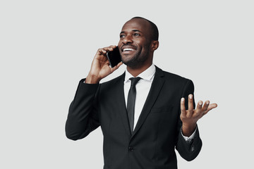 Happy young African man in formalwear talking on the smart phone and smiling while standing against...
