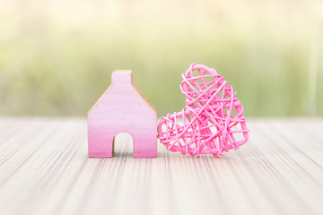 Pink wooden heart with miniature pink wooden house over blurred green garden background, outdoor day light, property and home business, house insurance and care