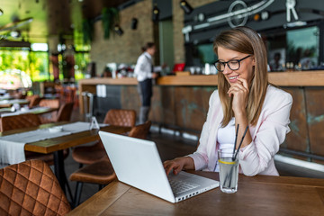 Young woman typing on laptop in cafe. Closeup portrait of smiling young beautiful woman leaning head on hand and working on laptop computer at cafe table