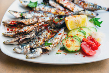 Fresh Grilled sardines served with fresh tomatoes, cucumbers slices and lemon on white plate for mediterranean healthy meal