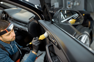Car service worker in uniform provides a professional vehicle interior cleaning, wiping door panel with a brush at the car service station