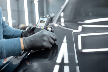 Inspector measuring paint cover thickness of the car body at the vehicle service box, close-up view on hands and professional thickness meter