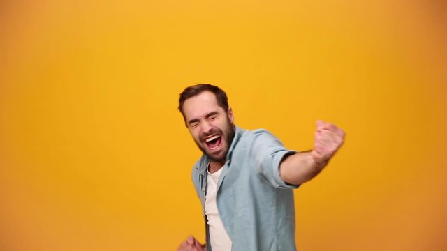 Handsome bearded young guy 20s in denim shirt white t-shirt isolated over yellow background in studio. People sincere emotions, lifestyle concept. Looking at the camera doing winner gesture, say Yeah