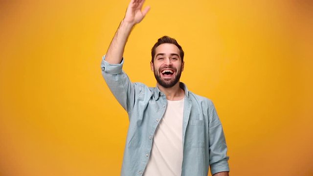Handsome bearded young guy 20s in denim shirt white t-shirt waving and greeting with hand as notices someone isolated over yellow background in studio. People sincere emotions, lifestyle concept.