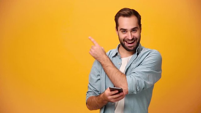 Handsome unshaven bearded young guy 20s in denim shirt white t-shirt isolated on yellow background in studio. People sincere emotions lifestyle concept. Pointing aside on copy space using mobile phone