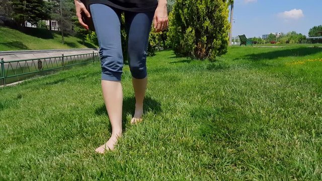 Front view of woman legs in blue tights walking slowly on barefoot on green grass in a park 