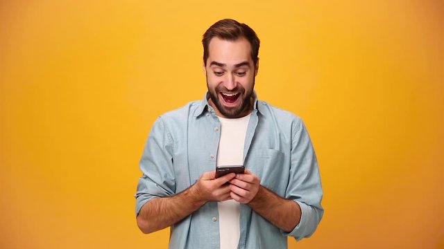 Handsome unshaven bearded young guy 20s in denim shirt white t-shirt isolated on yellow background in studio. People sincere emotions, lifestyle concept. Looking surprised wow, using mobile cell phone