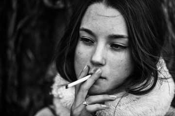 Smoking teenager in real life. Young beautiful white teeage girl in scarf smokes an cigarette near the tree on the street in the autumn. Deadly bad habit. Black and white.