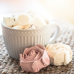 A cup of cocoa or chocolate next to a rose-shaped marshmallow. Warm cozy square background.