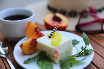 On a wooden table is a cup of coffee and a white plate with dessert and fruit. Hat and glasses are out of focus in the background. Close-up, horizontal, side view, cropped shot.