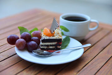 On a wooden table is a cup of coffee and a white plate with dessert and fruit. A piece of chocolate cake with white cream on a plate in a cafe. Close-up, horizontal, side view, cropped shot.