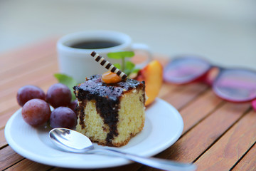 On a wooden table is a cup of coffee and a white plate with dessert and fruit. A piece of sponge cake on a white plate in a cafe. Close-up, horizontal, side view, cropped shot.