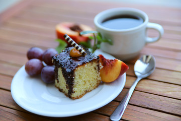 A piece of cake on a white plate. On a wooden table is a cup of coffee and a white plate with dessert and fruit. Close-up, horizontal, side view.
