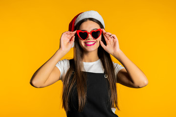 Beautiful woman with long hair in Santa hat and red heart-shaped sunglasses on studio yellow background.